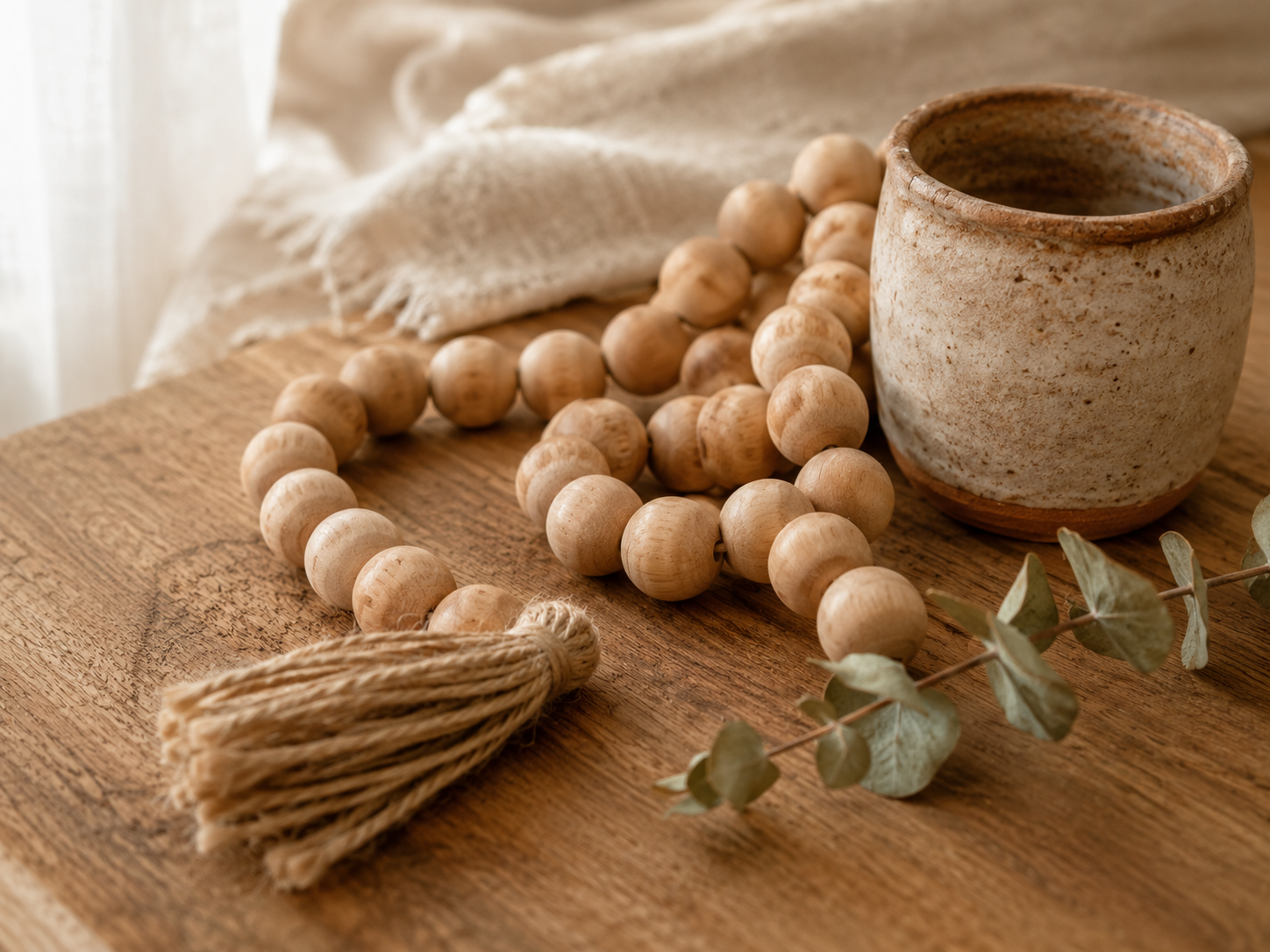 Close-up of a natural wood bead garland pooled on a warm wood surface beside a handmade ceramic pot and dried eucalyptus