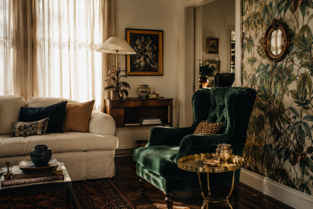 Editorial-style living room featuring an emerald velvet wing chair beside a linen sofa, a vintage brass side table, botanical wallpaper on one accent wall, and warm afternoon light filtering through sheer curtains.