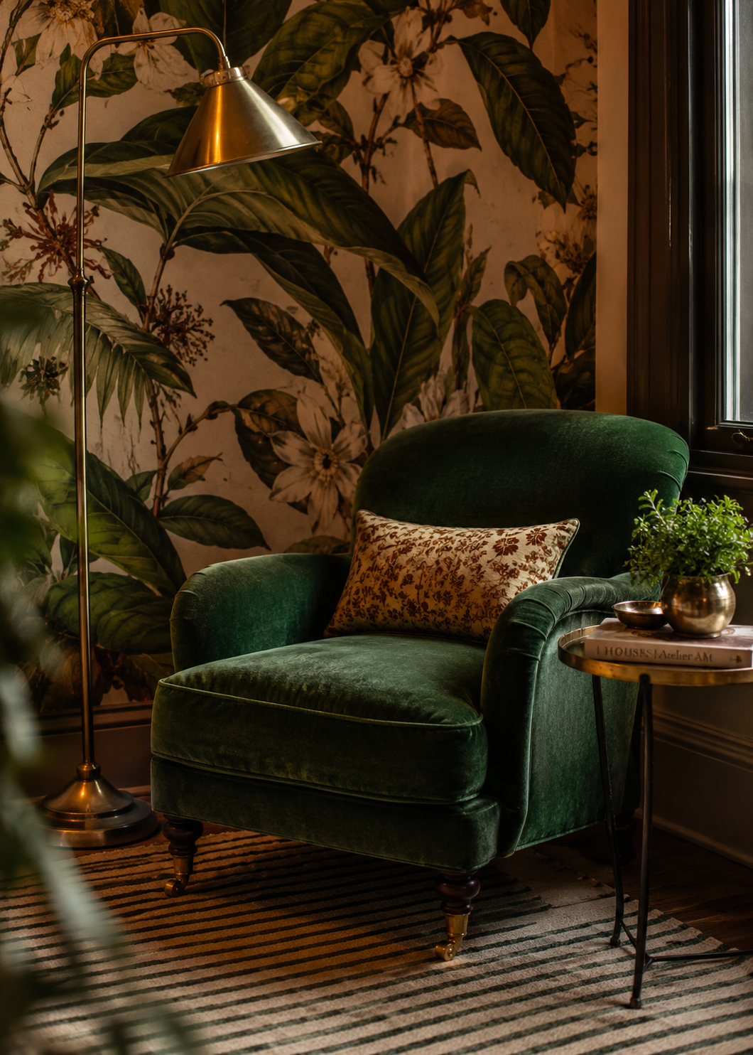 A reading nook corner layering a large-scale botanical wallpaper with a small-scale striped rug and a solid velvet armchair